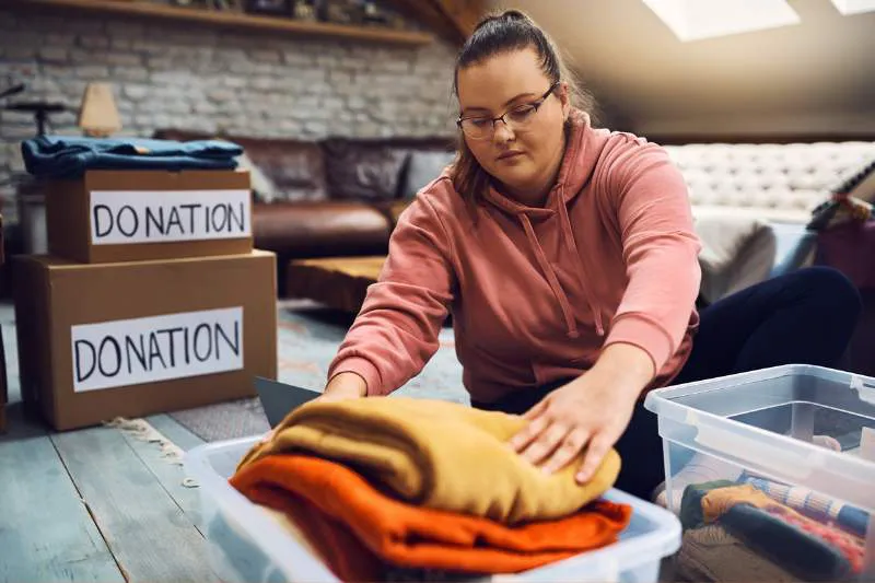 Woman folding clothes next to donation boxes, preparing items for charitable contributions to claim a tax deduction.